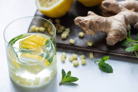selective focus of fresh ginger lemonade in glass with lemon and mint on wooden board on white...