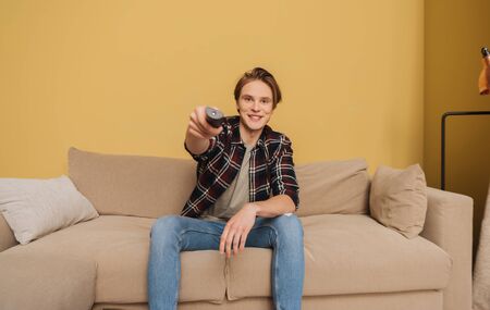 young smiling man holding remote controller and sitting on sofa, end of quarantine concept