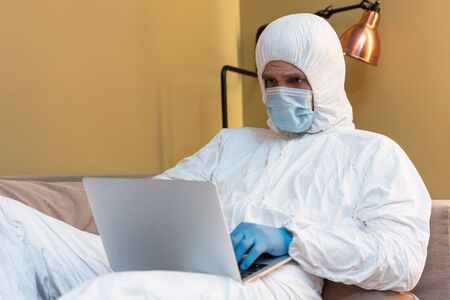 Selective focus of man in medical mask and hazmat suit using laptop on couchの写真素材