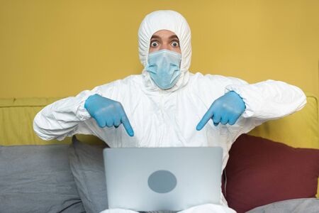 Shocked man in hazmat suit pointing with fingers on laptop on bed