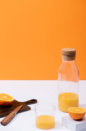 fresh orange juice in glass and bottle near cut fruit on wooden cutting board with knife on white surface isolated on orangeの写真素材
