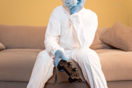 KYIV, UKRAINE - APRIL 24, 2020: Selective focus of man in medical mask, latex gloves and hazmat suit holding joystick on couchの写真素材