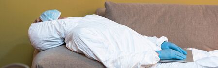 Panoramic shot of man in medical mask and hazmat suit holding laptop on couch
