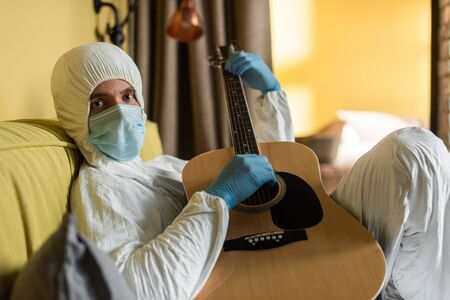 Selective focus of man in hazmat suit and medical mask looking at camera while playing acoustic...