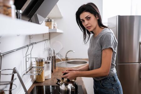 selective focus of woman with bruise on face touching teapot on electric stove in kitchen, domestic violence conceptの写真素材