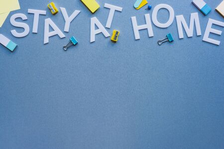 Top view of stay at home lettering near pencil sharpeners, erasers and binder clips on blue surface
