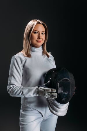 Attractive fencer in fencing suit holding mask and looking at camera isolated on black