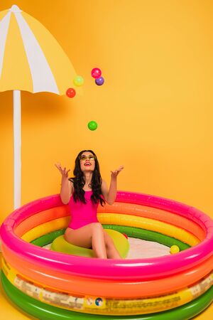 happy girl throwing in air balls while sitting in inflatable pool on yellow