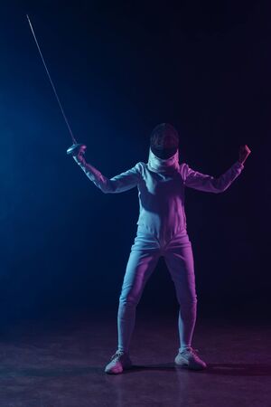Fencer showing yeah gesture while holding rapier on black background with lighting