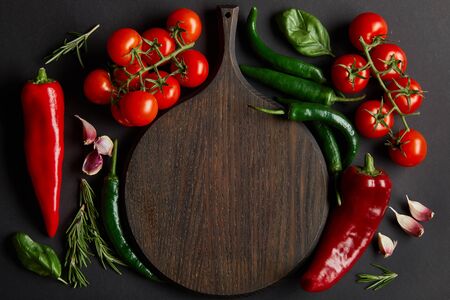 top view of wooden chopping board near ripe cherry tomatoes, garlic cloves, rosemary, basil leaves and green chili peppers on blackの写真素材