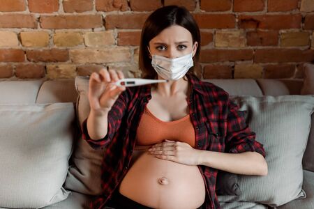 selective focus of pregnant woman in medical mask holding digital thermometer