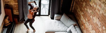 panoramic shot of cheerful and pregnant woman in wireless headphones dancing and holding remote...