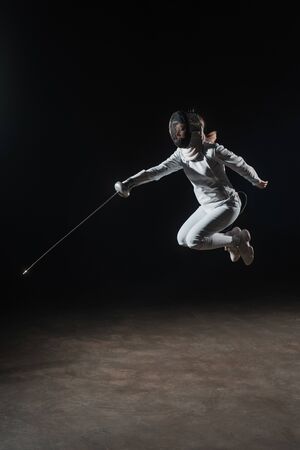 Fencer in fencing mask holding rapier and jumping on black backgroundの写真素材