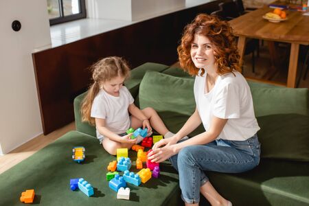 curly mother smiling near daughter playing building blocks in living roomの写真素材