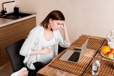 pregnant freelancer touching belly near laptop, smartphone, glass of water and fruits on tableの写真素材