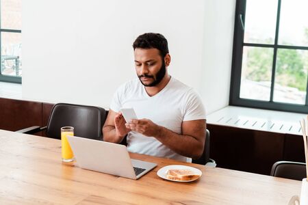 african american freelancer looking at smartphone near laptop near breakfast on tableの写真素材