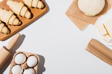top view of fresh croissants on wooden cutting board near raw dough, rolling pin, butter and eggs on...