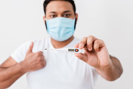 selective focus of african american man in medical mask holding digital thermometer and showing thumb up near white wallの写真素材