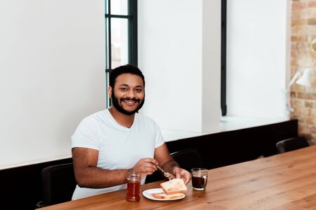 cheerful african american man holding spoon near jar with sweet jam and toast bread on plateの写真素材