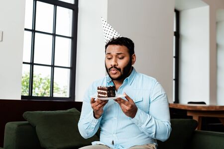 bearded african american man in party cap blowing out candle on birthday cakeの写真素材