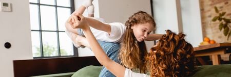 panoramic concept of curly mother lying on sofa, lifting happy daughter and holding hands in living roomの写真素材