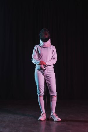Fencer in fencing suit and mask holding rapier on black background