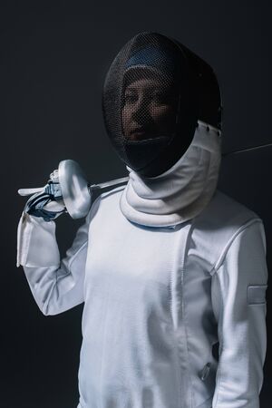 Fencer in fencing mask looking at camera and holding rapier isolated on blackの写真素材