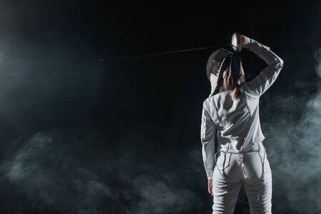 Back view of fencer holding rapier above head on black background with smoke
