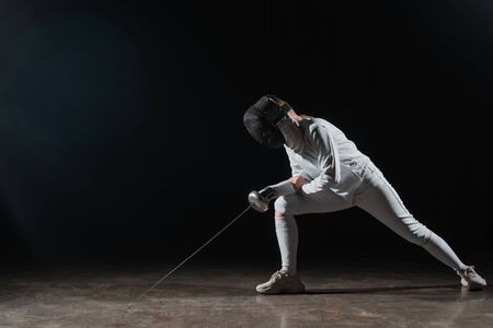 Fencer in fencing mask and suit holding rapier and doing lunge on black backgroundの写真素材