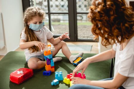 selective focus of cute kid in medical mask playing building blocks with redhead motherの写真素材