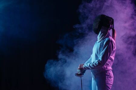 Side view of fencer in fencing mask holding rapier on black background with smoke