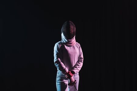 Fencer in fencing suit and mask holding rapier isolated on black