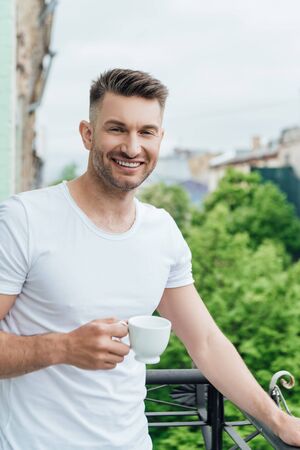 Handsome man smiling at camera while holding cup of coffee on balconyの写真素材