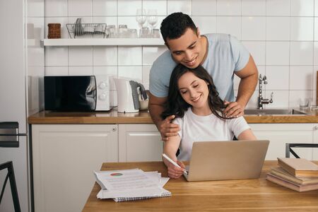 happy mixed race man hugging cheerful girlfriend and looking at laptopの写真素材