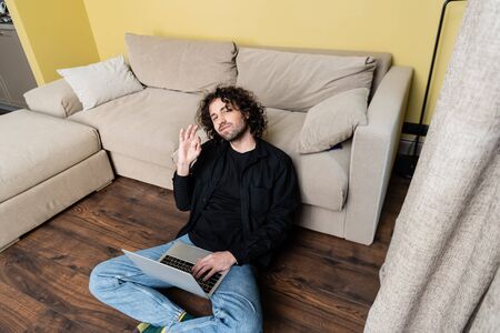 Selective focus of handsome man showing ok gesture while using laptop on floorの写真素材