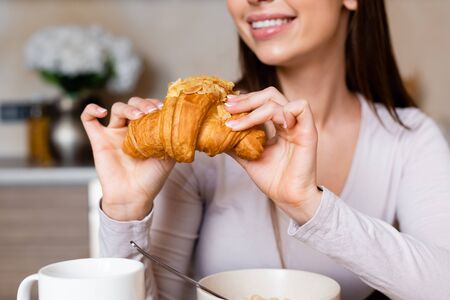 cropped view of happy girl holding tasty croissant near bowl and cupの写真素材