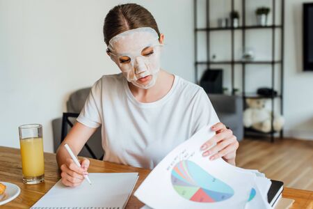 Selective focus of woman in face mask writing on notebook and holding papers with graphs near orange juice on tableの写真素材