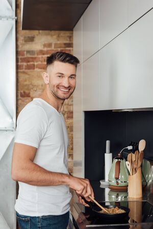Side view of handsome man smiling at camera while cooking noodles in frying pan in kitchenの写真素材
