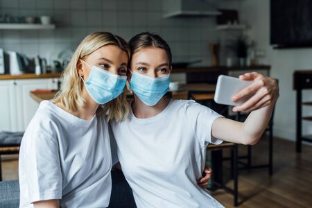 Sisters in medical masks taking selfie with smartphone at homeの写真素材
