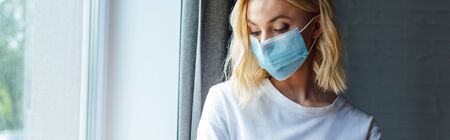 panoramic shot of young woman in medical mask standing near window at homeの写真素材