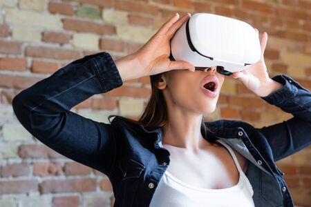 shocked young woman touching virtual reality headset against brick wall