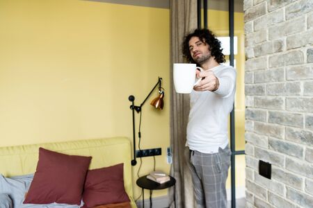 curly young man in pajamas holding cup of coffee in outstretched hand while standing in bedroomの写真素材