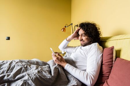 shocked young man touching hair while chatting on smartphone in bed