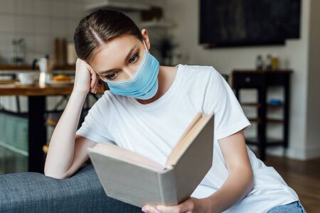 Selective focus of young woman in medical mask reading book in living roomの写真素材