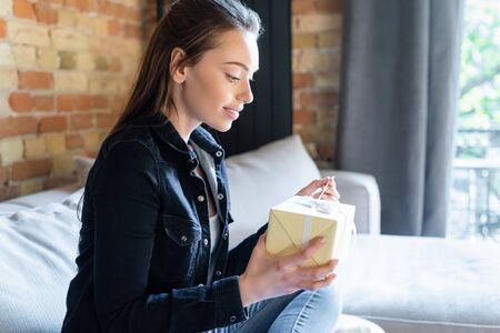side view of happy woman sitting on sofa and touching ribbon on gift boxの写真素材