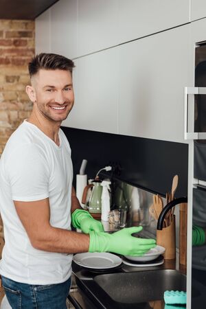 cheerful man wearing rubber gloves near white plates in kitchenの写真素材