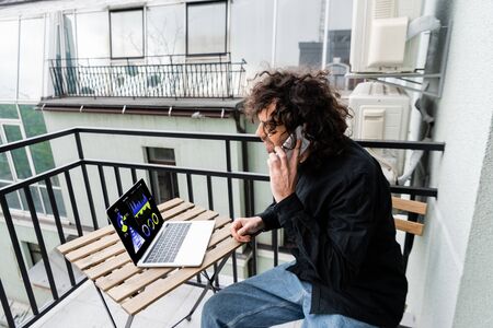 Man talking on smartphone near laptop with charts on table on balconyの写真素材