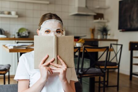 Young woman in face mask looking at camera while holding book at homeの写真素材
