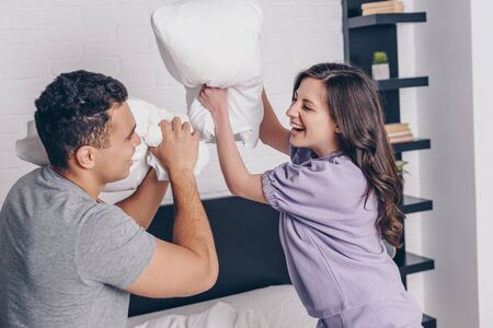cheerful interracial couple pillow fighting in bedroomの写真素材