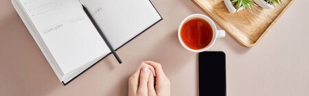 top view of female hands, smartphone near green plants on wooden board, cup of tea, planner with pencil on beige surface, panoramic shotの写真素材